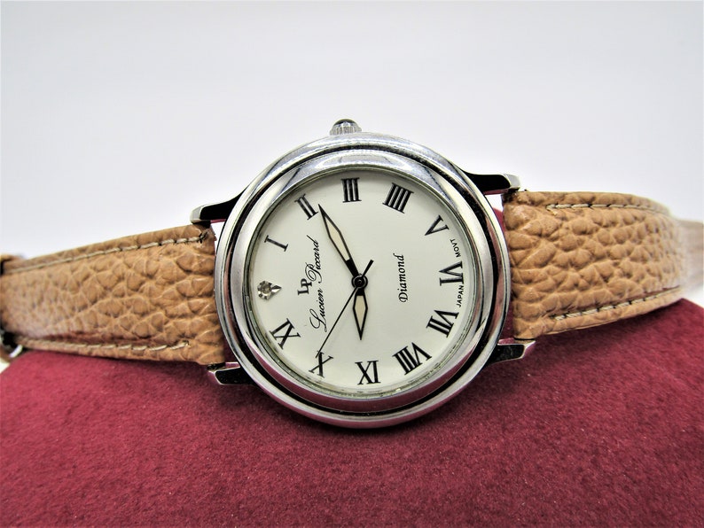 May include: A silver wristwatch with a white face and Roman numeral hour markers. The watch has a brown leather band and says "L.P. Lucien Piccard Diamond" on the face.