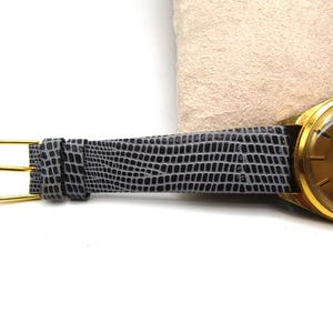 K&ouml;nnte beinhalten: Eine goldfarbene Armbanduhr mit einem grauen Echsenlederarmband. Das Zifferblatt der Uhr hat eine goldfarbene L&uuml;nette und ein wei&szlig;es Zifferblatt mit schwarzen Markierungen.