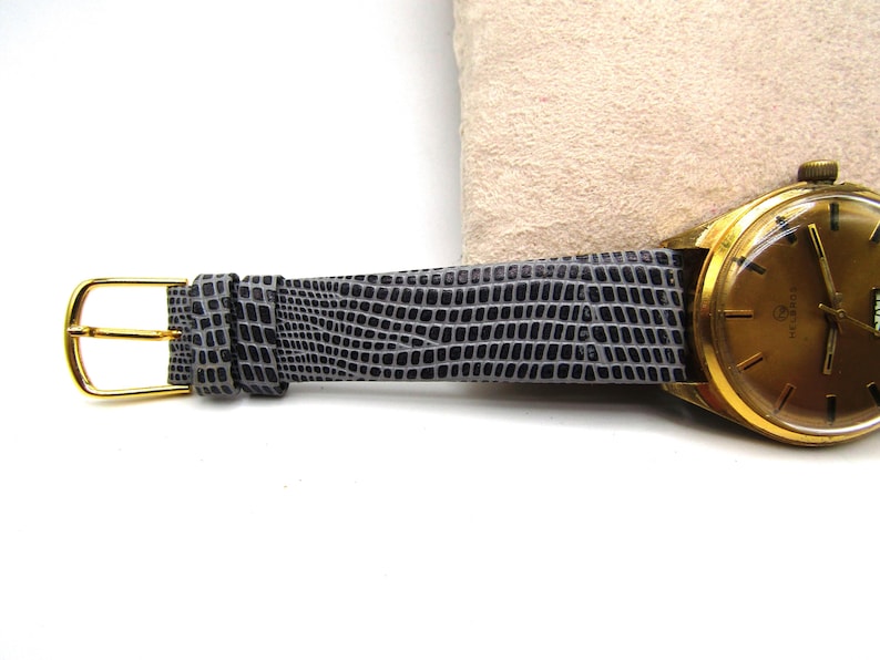 K&ouml;nnte beinhalten: Eine goldfarbene Armbanduhr mit einem grauen Echsenlederarmband. Das Zifferblatt der Uhr hat eine goldfarbene L&uuml;nette und ein wei&szlig;es Zifferblatt mit schwarzen Markierungen.