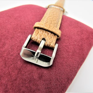 May include: A close-up of a brown leather watch strap with a silver buckle. The strap is on a burgundy velvet surface.