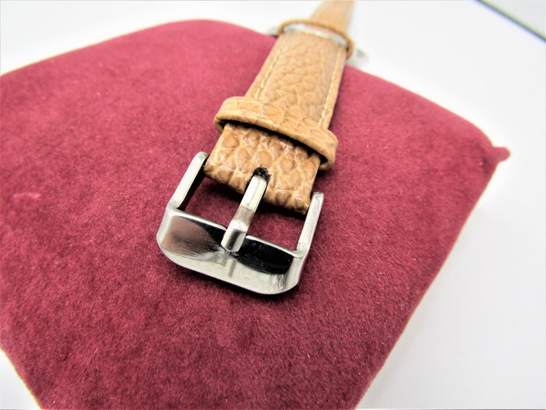 May include: A close-up of a brown leather watch strap with a silver buckle. The strap is on a burgundy velvet surface.