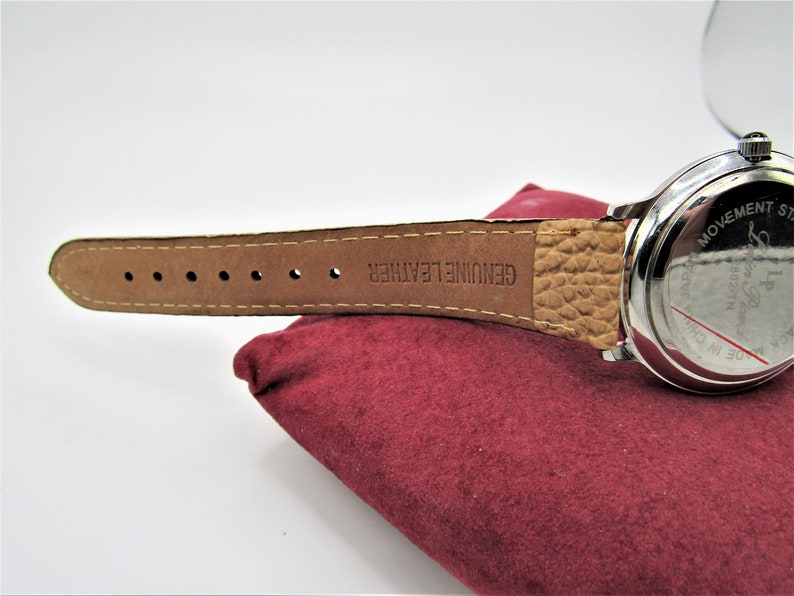 May include: A silver wristwatch with a brown leather band. The band is embossed with the words "GENUINE LEATHER". The back of the watch has the words "MADE IN CHINA" and "LP DESIGN" printed on it.