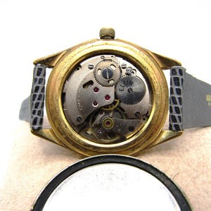 K&ouml;nnte beinhalten: Goldfarbene Armbanduhr mit schwarzem Lederarmband. Die R&uuml;ckseite der Uhr ist offen und zeigt das komplizierte Uhrwerk. Das Zifferblatt der Uhr tr&auml;gt die Aufschrift "ELGIN".