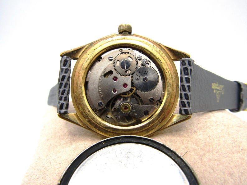 K&ouml;nnte beinhalten: Goldfarbene Armbanduhr mit schwarzem Lederarmband. Die R&uuml;ckseite der Uhr ist offen und zeigt das komplizierte Uhrwerk. Das Zifferblatt der Uhr tr&auml;gt die Aufschrift "ELGIN".