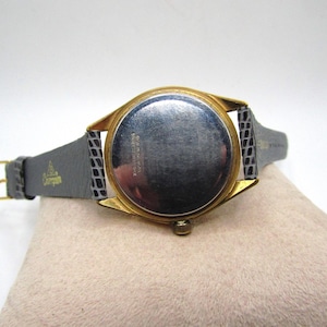 K&ouml;nnte beinhalten: Eine goldfarbene Armbanduhr mit einem schwarzen Lederarmband. Die Uhr hat ein rundes Zifferblatt und ein silbernes Zifferblatt. Die R&uuml;ckseite der Uhr ist sichtbar und zeigt die Aufschrift "Swiss Made" und andere Markierungen.