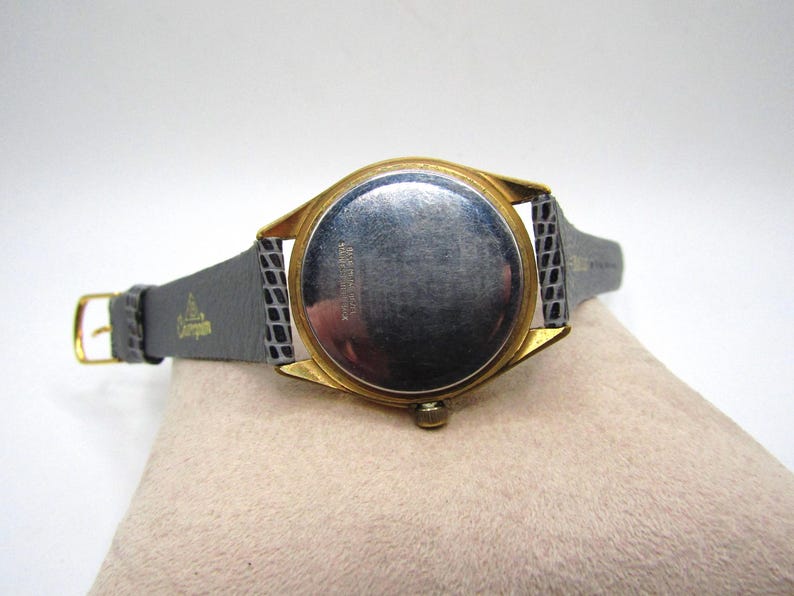 K&ouml;nnte beinhalten: Eine goldfarbene Armbanduhr mit einem schwarzen Lederarmband. Die Uhr hat ein rundes Zifferblatt und ein silbernes Zifferblatt. Die R&uuml;ckseite der Uhr ist sichtbar und zeigt die Aufschrift "Swiss Made" und andere Markierungen.