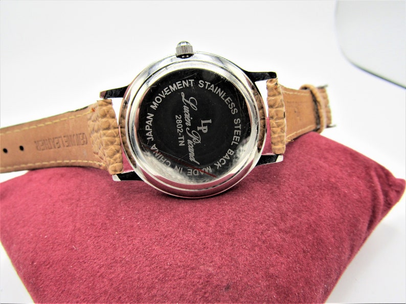 May include: Back of a silver wristwatch with a brown leather band. The watch face says "LP Lucien Piccard 2802-TN Made in China Japan Movement Stainless Steel Back".