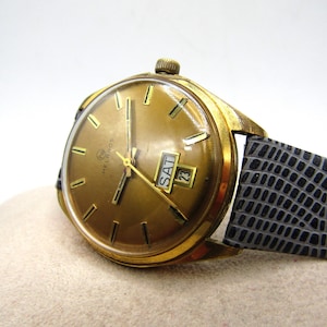 K&ouml;nnte beinhalten: Eine goldfarbene Armbanduhr mit einem schwarzen Lederarmband. Das Zifferblatt der Uhr hat einen goldenen Hintergrund mit schwarzen Stundenmarkierungen und Zeigern. Die Uhr hat ein Datumsfenster und ein Wochentagsfenster.