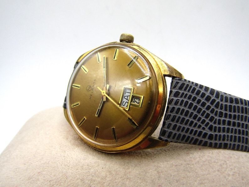K&ouml;nnte beinhalten: Eine goldfarbene Armbanduhr mit einem schwarzen Lederarmband. Das Zifferblatt der Uhr hat einen goldenen Hintergrund mit schwarzen Stundenmarkierungen und Zeigern. Die Uhr hat ein Datumsfenster und ein Wochentagsfenster.