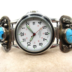 Puede incluir: Un reloj plateado con una esfera blanca y detalles negros. El reloj tiene una correa turquesa y plateada con un diseño decorativo. La esfera del reloj tiene el texto "100FT WATER RESIST QUARTZ" y "JAPAN MOVT".