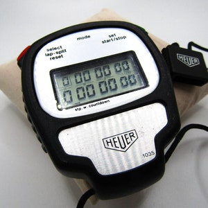 Puede incluir: Un cronómetro digital negro con una cara plateada y una correa negra. El cronómetro tiene una gran pantalla digital con el texto "stp.w. countdown" debajo. El cronómetro también tiene botones para "mode", "select lap-split reset", "set start/stop", y un logotipo HEUER en la cara. El cronómetro está sobre una superficie blanca.