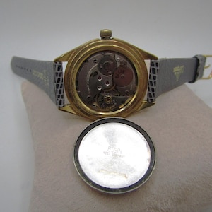 K&ouml;nnte beinhalten: Eine goldfarbene Armbanduhr mit einem grauen Lederarmband. Das Zifferblatt der Uhr ist freigelegt und zeigt das Innenleben der Uhr. Die R&uuml;ckseite des Uhrengeh&auml;uses ist mit dem Text "ELGIN WEST GERMANY" graviert.