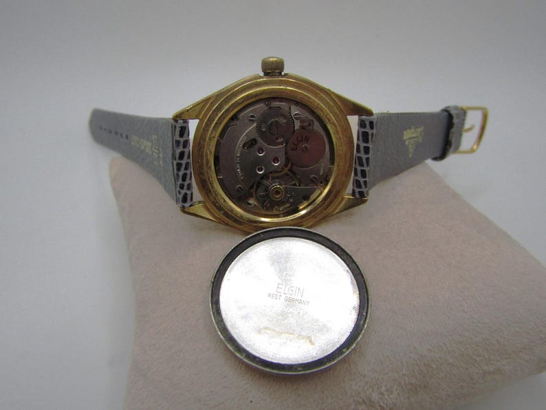 K&ouml;nnte beinhalten: Eine goldfarbene Armbanduhr mit einem grauen Lederarmband. Das Zifferblatt der Uhr ist freigelegt und zeigt das Innenleben der Uhr. Die R&uuml;ckseite des Uhrengeh&auml;uses ist mit dem Text "ELGIN WEST GERMANY" graviert.