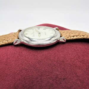 May include: A silver watch with a white face and Roman numeral hour markers. The watch has a brown leather band and is sitting on a red velvet surface.