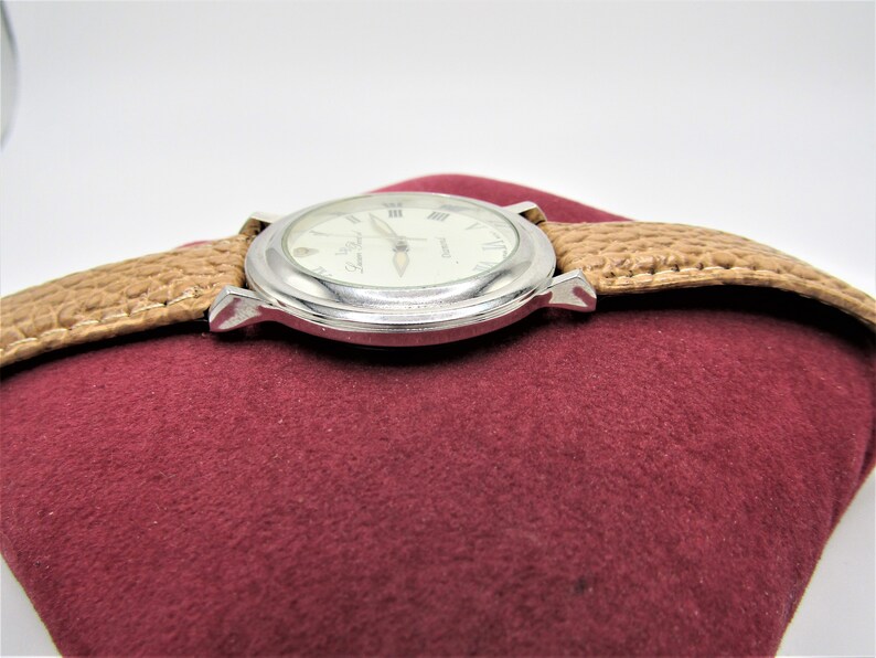 May include: A silver watch with a white face and Roman numeral hour markers. The watch has a brown leather band and is sitting on a red velvet surface.