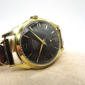 May include: A gold-tone wristwatch with a black face and gold-tone hands. The watch has a date window at the 3 o'clock position and the words "Geneva Luxe" printed on the face. The watch is on a gold-tone metal band.
