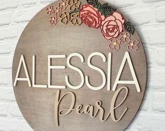 Puede incluir: Un letrero redondo de madera con el nombre "Alessia Pearl" en letras blancas y doradas. El letrero está decorado con una corona de flores rosas y amarillas.
