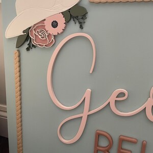Rectangular wooden nursery name sign featuring 3D lettering in blush pink and rose gold with floral cowgirl details. A white cowboy hat, boots, and rope frame add Western charm, while layered flowers in soft pink, cream, and sage green bring a feminine touch. Set against a pale blue background, this handcrafted wall art makes a unique personalized baby gift or statement piece for a floral Western-themed nursery.