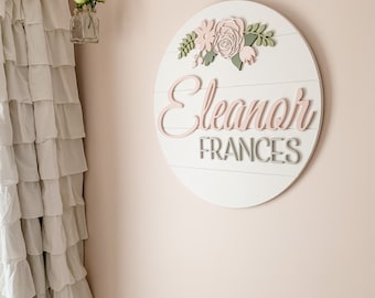 Puede incluir: Un letrero redondo de madera blanca con un diseño floral rosa y verde. El letrero dice "Eleanor Frances" en letras rosas y grises.
