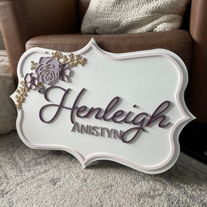 May include: White wooden sign with a pink and gold floral design and the name "Henleigh Anistyn" in purple lettering.