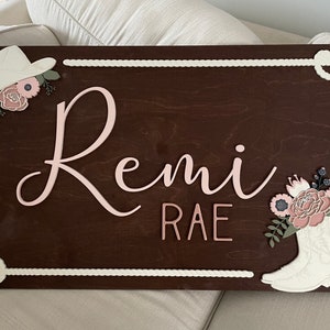 Rectangular wooden nursery name sign featuring 3D lettering in blush pink and rose gold with floral cowgirl details. A white cowboy hat, boots, and rope frame add Western charm, while layered flowers in soft pink, cream, and sage green bring a feminine touch. Set against a pale blue background, this handcrafted wall art makes a unique personalized baby gift or statement piece for a floral Western-themed nursery.