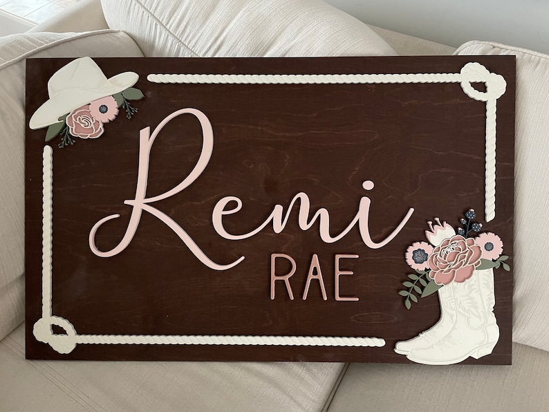 Rectangular wooden nursery name sign featuring 3D lettering in blush pink and rose gold with floral cowgirl details. A white cowboy hat, boots, and rope frame add Western charm, while layered flowers in soft pink, cream, and sage green bring a feminine touch. Set against a pale blue background, this handcrafted wall art makes a unique personalized baby gift or statement piece for a floral Western-themed nursery.
