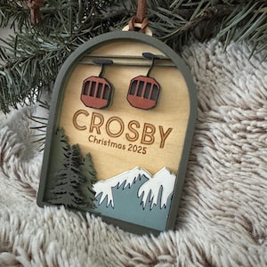 May include: A wooden Christmas decoration featuring a ski lift design. The ornament has the name "CROSBY" and "Christmas 2025" printed on it. It includes a brown leather strap for hanging and is set against a soft, textured background.