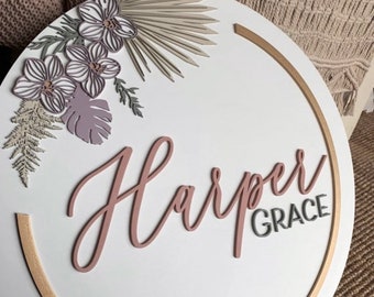 Puede incluir: Un letrero redondo de madera blanca con un círculo dorado alrededor del borde. El letrero tiene un diseño floral en tonos rosa y beige con el nombre "Harper Grace" escrito en cursiva.
