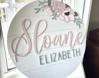 Puede incluir: Un letrero redondo de madera blanco con un diseño floral rosa y verde. El nombre "Sloane Elizabeth" está escrito en letras rosas y verdes.