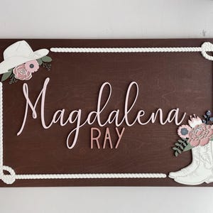 Rectangular wooden nursery name sign featuring 3D lettering in blush pink and rose gold with floral cowgirl details. A white cowboy hat, boots, and rope frame add Western charm, while layered flowers in soft pink, cream, and sage green bring a feminine touch. Set against a pale blue background, this handcrafted wall art makes a unique personalized baby gift or statement piece for a floral Western-themed nursery.