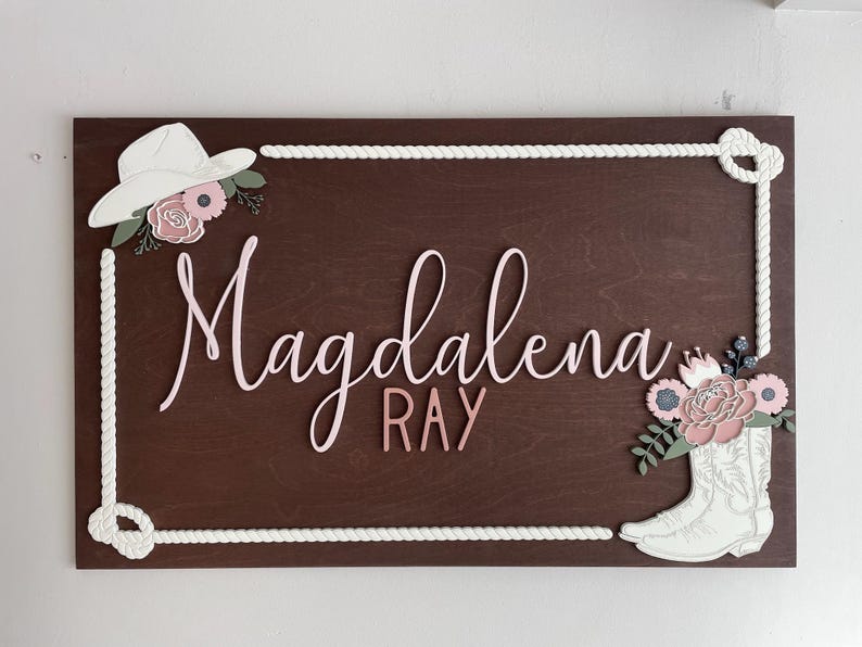 Rectangular wooden nursery name sign featuring 3D lettering in blush pink and rose gold with floral cowgirl details. A white cowboy hat, boots, and rope frame add Western charm, while layered flowers in soft pink, cream, and sage green bring a feminine touch. Set against a pale blue background, this handcrafted wall art makes a unique personalized baby gift or statement piece for a floral Western-themed nursery.
