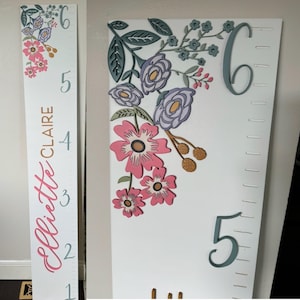 May include: White wooden height chart with floral decorations in pink, purple, and gold. The chart has the name "Ellietie Claire" in pink script, with measurements up to 183 cm. Numbers and markings are in a soft blue.