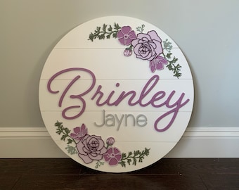 Puede incluir: Un letrero redondo de madera blanca con los nombres "Brinkley Jayne" escritos en letras cursivas moradas. El letrero está decorado con una corona de flores moradas y verdes.