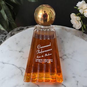 Puede incluir: Un frasco de perfume vintage "Command Performance" de Helena Rubinstein. El perfume de color ámbar está en un frasco acanalado en forma de trapecio con un tapón esférico dorado. La etiqueta dice "Eau de Parfum" y "4 fl oz."