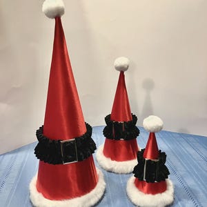 Puede incluir: Tres gorros de Papá Noel rojos con ribete de piel blanca y cinturones de terciopelo negro. Los gorros son de diferentes tamaños, siendo el más grande el del centro.