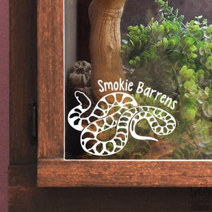 May include: A white vinyl decal applied to a glass surface featuring a stylised snake illustration with a patterned body and the text Smokie Barrens written in a white sans-serif font positioned above the snake graphic.