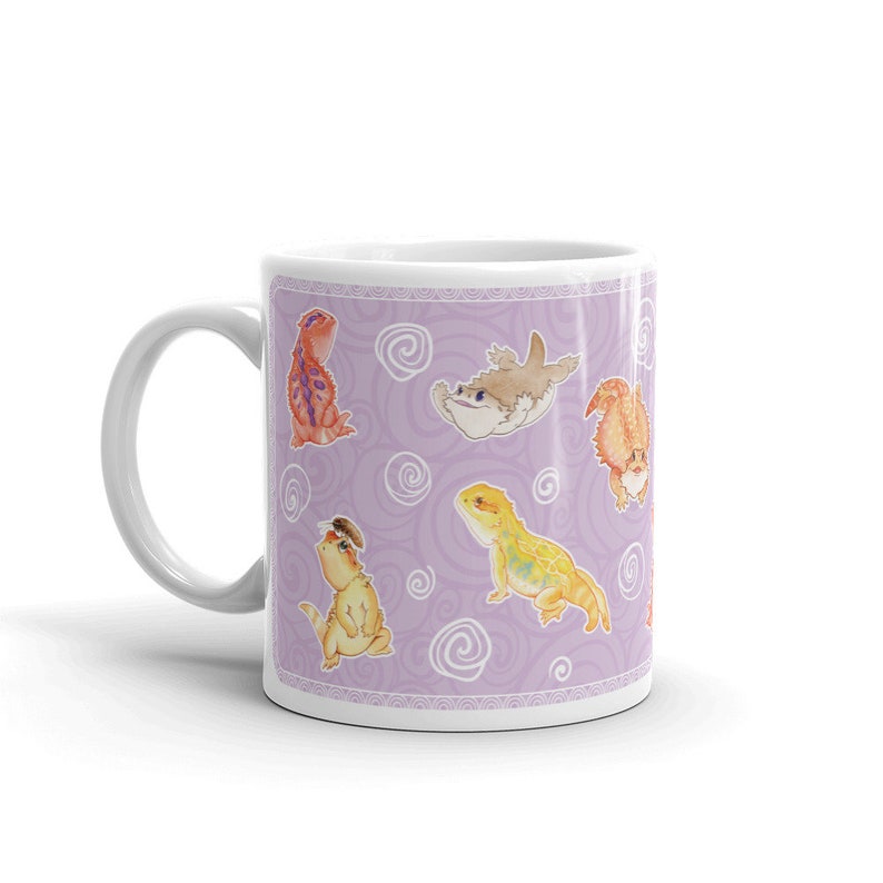 May include: A white ceramic mug with a purple background and a repeating pattern of cartoon bearded dragons and a fish. The mug has a white handle.
