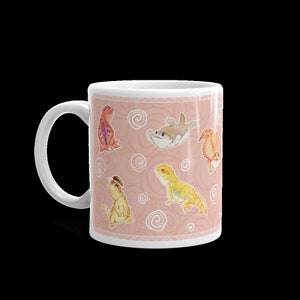 May include: White ceramic mug with a pink background and a repeating pattern of cartoon bearded dragons and a shark. The bearded dragons are in various poses and colors, including orange, yellow, and red.