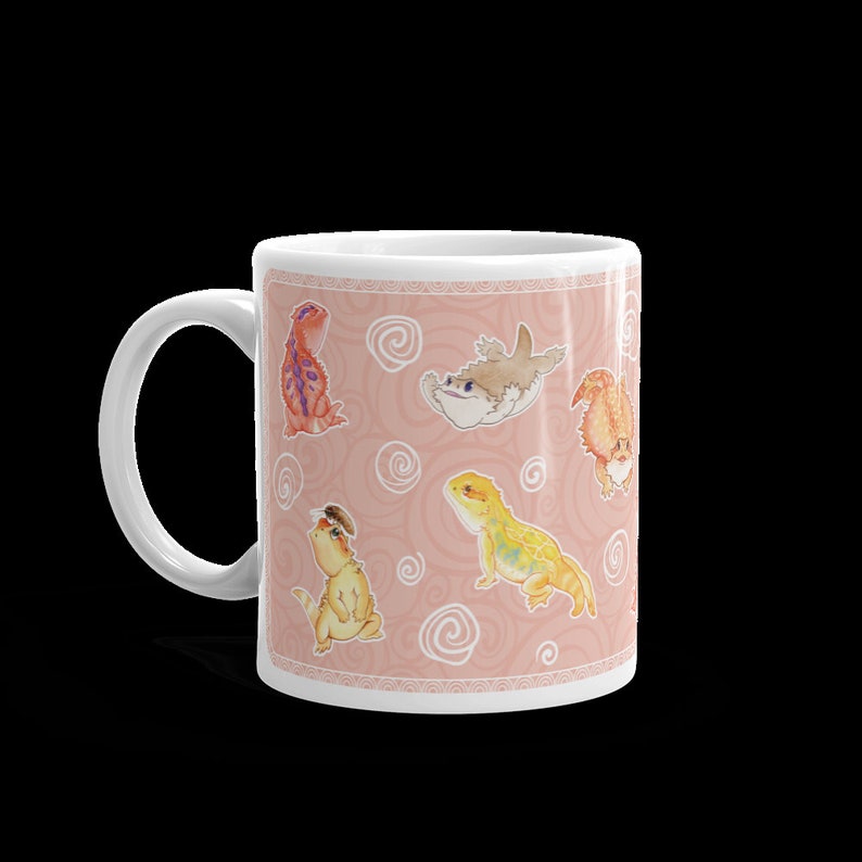 May include: White ceramic mug with a pink background and a repeating pattern of cartoon bearded dragons and a shark. The bearded dragons are in various poses and colors, including orange, yellow, and red.