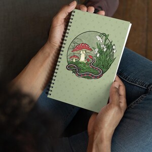 Könnte beinhalten: Ein spiralgebundenes Notizbuch mit einem hellen salbeigrünen Einband, der mit einem wiederkehrenden Muster aus kleinen Pilzsymbolen bedruckt ist. In der Mitte befindet sich eine kreisförmige Illustration mit rotköpfigen Pilzen, weißen Glockenblumen und einer schwarz-roten Schlange.