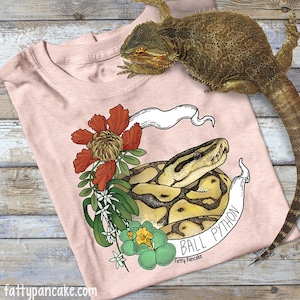 May include: A pink t-shirt featuring a graphic of a ball python snake with floral accents. The text "Ball Python" is written on a banner with the artist's name "Fatty Pancake" below it.