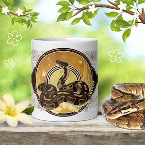 May include: A white ceramic mug featuring a circular graphic of a brown and black patterned ball python snake. The design includes a golden-yellow background with subtle line art of butterflies and tall grasses, centered on the mug's exterior surface.