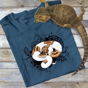 May include: A dark blue t-shirt featuring a white and brown snake design. The snake is curled up in a circle with its head facing the viewer. The design is surrounded by black swirls and splatters.