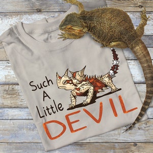 May include: A beige t-shirt featuring a cartoon illustration of a spiky, red and white lizard with the text "Such A Little Devil" printed in orange.