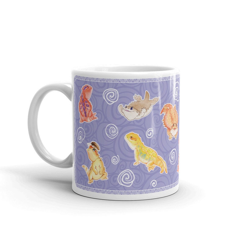 May include: A white ceramic mug with a purple background and a repeating pattern of cartoon bearded dragons and a fish. The bearded dragons are in various poses and colors, including orange, yellow, and green.