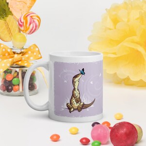 May include: A white ceramic mug featuring a graphic illustration of a brown spotted lizard sitting in grass with a small blue butterfly perched on its nose, set against a light purple background with faint white swirling line patterns.