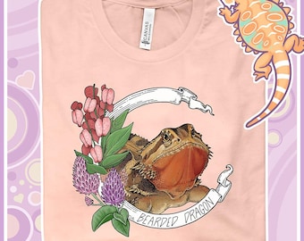 Può includere: Una maglietta rosa con una grafica di un drago barbuto circondato da fiori e uno striscione che recita "Bearded Dragon".