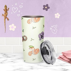 May include: A pale green stainless steel tumbler featuring a repeating pattern of cartoon snakes in orange, brown, and purple, with small green leaf accents. The tumbler includes a clear, removable plastic lid with a black sliding closure mechanism.