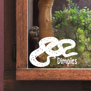 Peut inclure: Un autocollant en vinyle blanc représentant un serpent enroulé avec de petites taches noires, appliqué sur une surface en verre. Le mot "Dimples" est imprimé en lettres blanches sans empattement sous l'illustration du serpent.