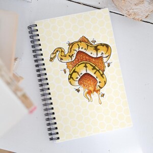 A spiral-bound notebook with a pale yellow honeycomb-patterned cover, featuring an illustration of a yellow and black snake wrapped in orange honeycomb with bees, photographed on a white surface.